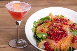 Una ensalada vibrante cubierta con frutos rojos complementa un cóctel rosa en una copa de martini, seduciendo a los comensales del Restaurante La Cocotte Cocina internacional en Málaga, todo elegantemente dispuesto en una encantadora mesa de madera. - ForTwo - Planes para parejas