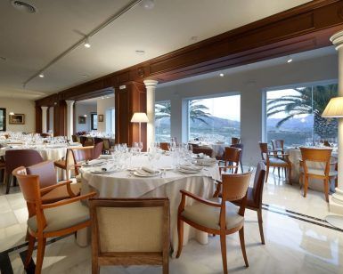 El Menú romántico en el restaurante Abades (Ronda) ofrece una experiencia gastronómica inolvidable con elegantes mesas redondas, sillas de madera, manteles blancos y grandes ventanales que muestran cautivadoras vistas a las palmeras. - ForTwo - Planes para parejas
