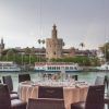 En el Restaurante Abades Triana de Sevilla, una mesa de lujo está dispuesta junto a una ventana con vistas al río, la torre histórica y los barcos. - ForTwo - Planes para parejas