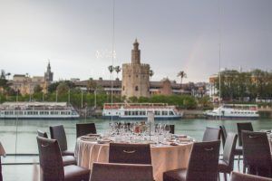 En el Restaurante Abades Triana de Sevilla, una mesa de lujo está dispuesta junto a una ventana con vistas al río, la torre histórica y los barcos. - ForTwo - Planes para parejas