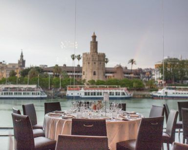 En el Restaurante Abades Triana de Sevilla, una mesa de lujo está dispuesta junto a una ventana con vistas al río, la torre histórica y los barcos. - ForTwo - Planes para parejas