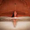 En la tranquila piscina rojiza del Hammam Al Ándalus, una persona se relaja mientras el agua cae en cascada sobre su cabeza. El Baño y ritual Alfolí canaliza la esencia relajante de las antiguas tradiciones en el centro de Madrid. - ForTwo - Planes para parejas