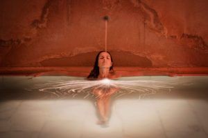 En la tranquila piscina rojiza del Hammam Al Ándalus, una persona se relaja mientras el agua cae en cascada sobre su cabeza. El Baño y ritual Alfolí canaliza la esencia relajante de las antiguas tradiciones en el centro de Madrid. - ForTwo - Planes para parejas