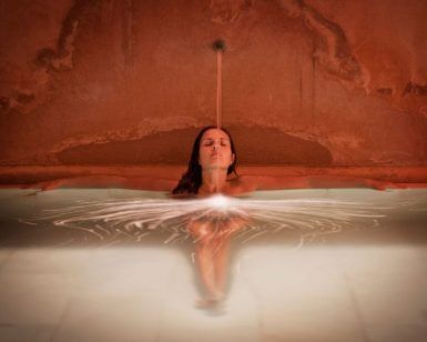 En la tranquila piscina rojiza del Hammam Al Ándalus, una persona se relaja mientras el agua cae en cascada sobre su cabeza. El Baño y ritual Alfolí canaliza la esencia relajante de las antiguas tradiciones en el centro de Madrid. - ForTwo - Planes para parejas