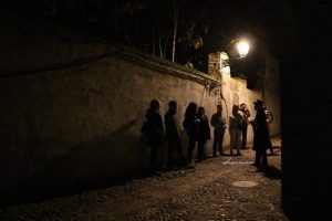 Un grupo se reúne en un callejón poco iluminado del Albaicín, iluminado por una farola, mientras su guía, con sombrero, comparte apasionadamente las historias ocultas de Granada en el Tour Conspiración en el Albaicín Granada. - ForTwo - Planes para parejas