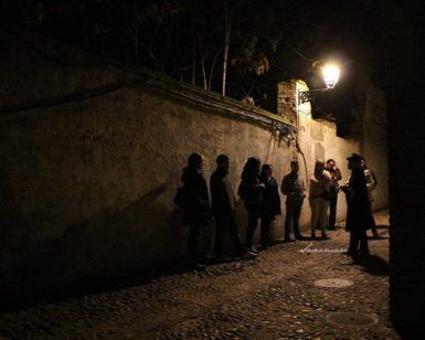 Un grupo se reúne en un callejón poco iluminado del Albaicín, iluminado por una farola, mientras su guía, con sombrero, comparte apasionadamente las historias ocultas de Granada en el Tour Conspiración en el Albaicín Granada. - ForTwo - Planes para parejas