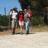 Tres excursionistas con mochilas posan felices en un camino de grava, abrazados por frondosos árboles y una cerca bajo el cielo azul claro, llenos del espíritu de "Senderismo de día y medio: Del Romero al Aguacate - Granada", mientras se deleitan en su viaje juntos. - ForTwo - Planes para parejas