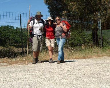 Tres excursionistas con mochilas posan felices en un camino de grava, abrazados por frondosos árboles y una cerca bajo el cielo azul claro, llenos del espíritu de "Senderismo de día y medio: Del Romero al Aguacate - Granada", mientras se deleitan en su viaje juntos. - ForTwo - Planes para parejas