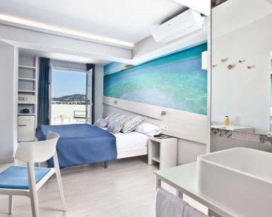 Disfrute de una habitación moderna en el Escapada Hotel Ryans La Marina con cama doble, detalles en azul, obras de arte playeras y balcón con vistas al mar. Disfrute del ambiente ibicenco con desayuno incluido para empezar el día. - ForTwo - Planes para parejas