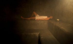 Envuelta en una toalla, la persona se sienta serenamente en un banco de piedra en la sala de vapor tenuemente iluminada de Rituels d'Orient Barcelona, abrazando el ambiente tranquilo que recuerda al Hammam Ritual de los Sultanes. - ForTwo - Planes para parejas