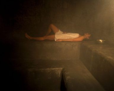 Envuelta en una toalla, la persona se sienta serenamente en un banco de piedra en la sala de vapor tenuemente iluminada de Rituels d'Orient Barcelona, abrazando el ambiente tranquilo que recuerda al Hammam Ritual de los Sultanes. - ForTwo - Planes para parejas