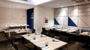 El comedor luce arte mural geométrico, mesas de madera y platos y vasos cuidadosamente colocados. Disfrute del sabor griego en el Restaurante Griego Kuzina en Valencia, donde la elegancia mediterránea se fusiona con el diseño contemporáneo para una comida inolvidable. - ForTwo - Planes para parejas