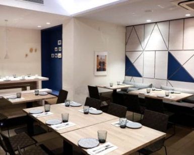 El comedor luce arte mural geométrico, mesas de madera y platos y vasos cuidadosamente colocados. Disfrute del sabor griego en el Restaurante Griego Kuzina en Valencia, donde la elegancia mediterránea se fusiona con el diseño contemporáneo para una comida inolvidable. - ForTwo - Planes para parejas