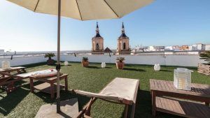 Disfruta de una tranquila mañana en la azotea con el desayuno de "Escapadita a San Fernando con desayuno, cava y bombones (Cádiz)". Relájate bajo una sombrilla entre tumbonas y exuberante vegetación, contemplando el vibrante paisaje urbano de Cádiz y las majestuosas torres de la iglesia de San Fernando a lo lejos. - ForTwo - Planes para parejas