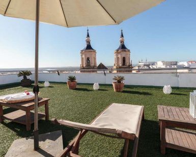 Disfruta de una tranquila mañana en la azotea con el desayuno de "Escapadita a San Fernando con desayuno, cava y bombones (Cádiz)". Relájate bajo una sombrilla entre tumbonas y exuberante vegetación, contemplando el vibrante paisaje urbano de Cádiz y las majestuosas torres de la iglesia de San Fernando a lo lejos. - ForTwo - Planes para parejas