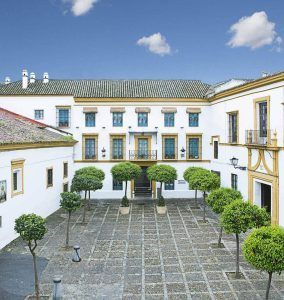 El patio del Hotel de 4 estrellas Hospes Las Casas del Rey Baeza en Sevilla cuenta con árboles simétricos, un techo de tejas y un cielo azul claro, creando un oasis de relajación. - ForTwo - Planes para parejas