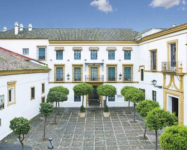 El patio del Hotel de 4 estrellas Hospes Las Casas del Rey Baeza en Sevilla cuenta con árboles simétricos, un techo de tejas y un cielo azul claro, creando un oasis de relajación. - ForTwo - Planes para parejas