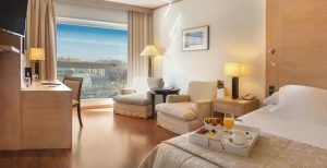 La encantadora habitación del Hotel SH Valencia Palace (Valencia) incluye una cómoda cama, elegantes sillas y una mesa junto a la ventana con vistas a la ciudad. Una bandeja de desayuno repleta de deliciosos dulces le espera en la cama. - ForTwo - Planes para parejas