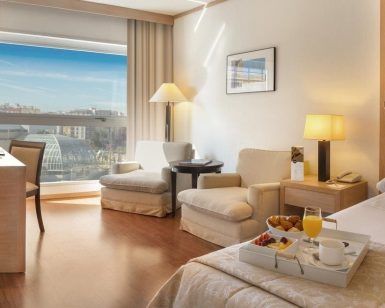 La encantadora habitación del Hotel SH Valencia Palace (Valencia) incluye una cómoda cama, elegantes sillas y una mesa junto a la ventana con vistas a la ciudad. Una bandeja de desayuno repleta de deliciosos dulces le espera en la cama. - ForTwo - Planes para parejas