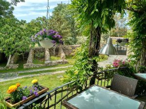 Mesas, plantas colgantes y flores crean un encantador patio con jardín contra una pared de piedra rústica, capturando la esencia tranquila de Almazara de San Pedro - Casa rural en Sierra de Gata con baño árabe (Cáceres), con exuberante vegetación para una escena encantadora. - ForTwo - Planes para parejas