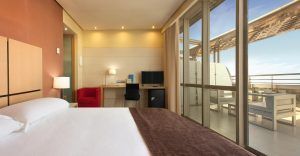 Alójese en una elegante habitación del Hotel Silken Puerta de Valencia con una cama grande, escritorio y silla roja. Disfrute de su café matutino en el balcón mientras disfruta de las soleadas vistas a través de las puertas de cristal, ideal para su visita. - ForTwo - Planes para parejas
