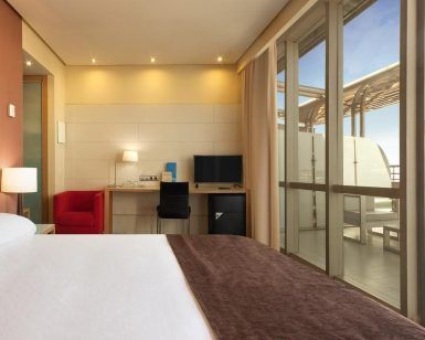 Alójese en una elegante habitación del Hotel Silken Puerta de Valencia con una cama grande, escritorio y silla roja. Disfrute de su café matutino en el balcón mientras disfruta de las soleadas vistas a través de las puertas de cristal, ideal para su visita. - ForTwo - Planes para parejas