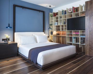 Crea un dormitorio moderno inspirado en el Hotel Sir Joan de Ibiza, con una pared con acento azul, pisos de madera, estantes elegantes y una cama acogedora vestida con sábanas blancas y una manta oscura. - ForTwo - Planes para parejas