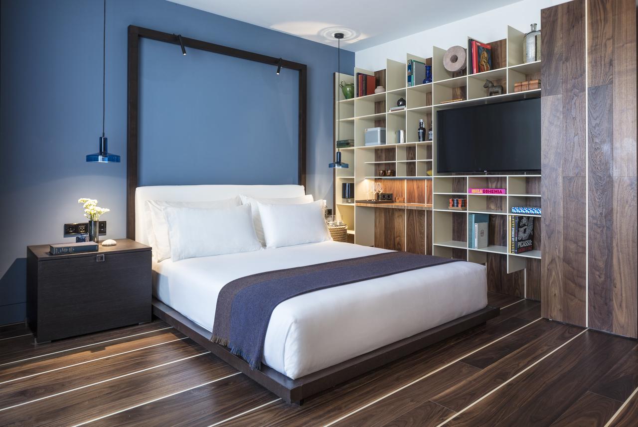 Crea un dormitorio moderno inspirado en el Hotel Sir Joan de Ibiza, con una pared con acento azul, pisos de madera, estantes elegantes y una cama acogedora vestida con sábanas blancas y una manta oscura. - ForTwo - Planes para parejas