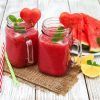 Dos frascos de batido de sandía con rodajas en forma de corazón, menta y pajitas descansan sobre una esterilla; cerca, una refrescante sandía y un limón. Ideal para una mañana en el Hotel Abades Nevada Palace de Granada, donde Viernes Spa + Smoothie ofrece relajación entre sabores vibrantes. - ForTwo - Planes para parejas