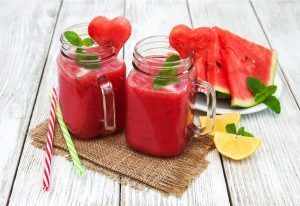 Dos frascos de batido de sandía con rodajas en forma de corazón, menta y pajitas descansan sobre una esterilla; cerca, una refrescante sandía y un limón. Ideal para una mañana en el Hotel Abades Nevada Palace de Granada, donde Viernes Spa + Smoothie ofrece relajación entre sabores vibrantes. - ForTwo - Planes para parejas