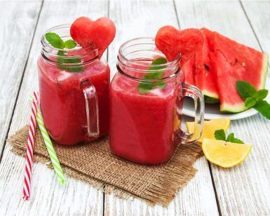 Dos frascos de batido de sandía con rodajas en forma de corazón, menta y pajitas descansan sobre una esterilla; cerca, una refrescante sandía y un limón. Ideal para una mañana en el Hotel Abades Nevada Palace de Granada, donde Viernes Spa + Smoothie ofrece relajación entre sabores vibrantes. - ForTwo - Planes para parejas