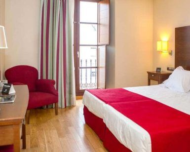Experimente la comodidad y el estilo en Escapada Hotel Soho Boutique Casa Don Fernando, Cáceres. Disfrute de una acogedora habitación con cama doble, manta roja, silla, escritorio, televisión y balcón con cortinas de rayas. Incluye desayuno, cata de ibéricos y vino en el corazón de la ciudad. - ForTwo - Planes para parejas
