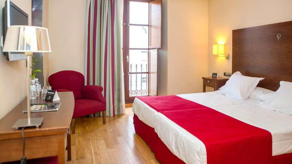 Experimente la comodidad y el estilo en Escapada Hotel Soho Boutique Casa Don Fernando, Cáceres. Disfrute de una acogedora habitación con cama doble, manta roja, silla, escritorio, televisión y balcón con cortinas de rayas. Incluye desayuno, cata de ibéricos y vino en el corazón de la ciudad. - ForTwo - Planes para parejas
