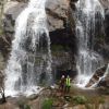 Cerca de la cascada en un acantilado rocoso, una pareja con cascos sonríe, abrazando la aventura de "Barranquismo en pareja en Somosierra - Madrid. - ForTwo - Planes para parejas