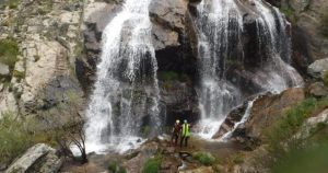 Cerca de la cascada en un acantilado rocoso, una pareja con cascos sonríe, abrazando la aventura de "Barranquismo en pareja en Somosierra - Madrid. - ForTwo - Planes para parejas