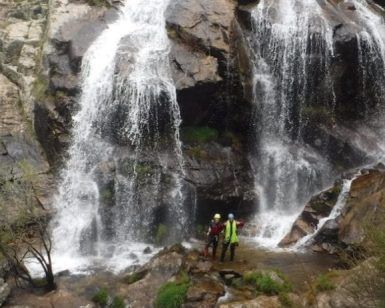 Cerca de la cascada en un acantilado rocoso, una pareja con cascos sonríe, abrazando la aventura de "Barranquismo en pareja en Somosierra - Madrid. - ForTwo - Planes para parejas
