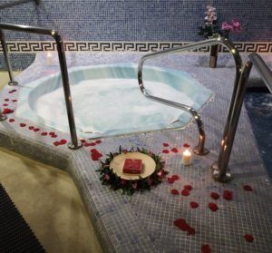 El Spa para 2 personas con masaje relajante opcional en Aquabody Wellness & SPA (Madrid) ofrece un lujoso jacuzzi para dos, adornado con pétalos de rosa y velas para un ambiente perfecto, complementado con un libro en una corona en esta serena sala de spa con azulejos. - ForTwo - Planes para parejas