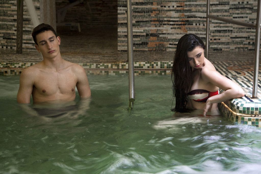 En Termas de Hispalis, una mujer se relaja en el borde de la piscina y un hombre se encuentra en agua tibia, ambos disfrutando de la relajante experiencia del Circuito Termal con Masaje. - ForTwo - Planes para parejas
