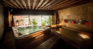 Una acogedora sala de spa con jacuzzi, iluminación ambiental y exuberante vegetación refleja el estilo del Spa y masaje a lo japonés + menú de tapas en Madrid. Faroles rojos decorativos en una pared de piedra aportan un toque elegante. - ForTwo - Planes para parejas
