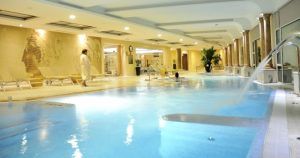 El Circuito Relax del Auditorio Beatriz Toledo cuenta con una tranquila piscina interior con tumbonas, una persona en bata y un nadador rodeados de una elegante iluminación y decoración. - ForTwo - Planes para parejas