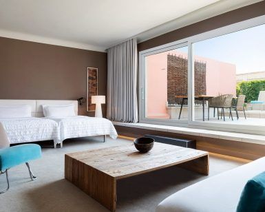 Descubra la comodidad de nuestra Suite Grand Terrace, la comodidad hecha habitación (Barcelona), con dos camas, una mesa de madera y una silla azul. Disfrute de la vista a través de un amplio ventanal que da a una terraza con acogedores asientos. - ForTwo - Planes para parejas