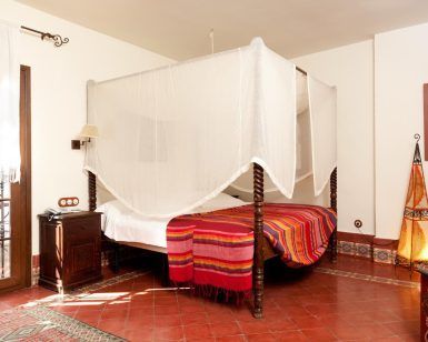 Un acogedor dormitorio con cama con dosel, manta colorida, muebles de madera y linterna decorativa sobre el suelo de baldosas rojas ofrece el ambiente perfecto para quienes buscan Suites románticas con jacuzzi en Alcoba del Rey de Sevilla (Sevilla). - ForTwo - Planes para parejas