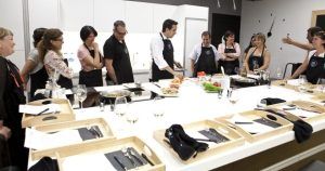 Personas con delantales observan a un chef en el mostrador de la cocina, mostrando habilidades culinarias durante una vibrante experiencia de Taller de Cocina en Illa Diagonal en Barcelona. - ForTwo - Planes para parejas
