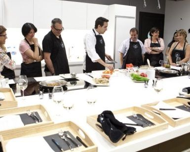 Personas con delantales observan a un chef en el mostrador de la cocina, mostrando habilidades culinarias durante una vibrante experiencia de Taller de Cocina en Illa Diagonal en Barcelona. - ForTwo - Planes para parejas