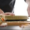 Primer plano de unas manos enrollando sushi con destreza con una estera de bambú, capturando la esencia de la auténtica preparación de sushi en el Taller de preparación de sushi (Madrid). - ForTwo - Planes para parejas