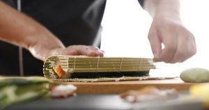 Primer plano de unas manos enrollando sushi con destreza con una estera de bambú, capturando la esencia de la auténtica preparación de sushi en el Taller de preparación de sushi (Madrid). - ForTwo - Planes para parejas