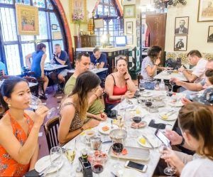 Gente disfrutando de un "Tour de tapas por Barcelona" en un animado restaurante, cenando en una mesa larga con bebidas, rodeados de una decoración colorida que recuerda a una vibrante noche de Barcelona. - ForTwo - Planes para parejas
