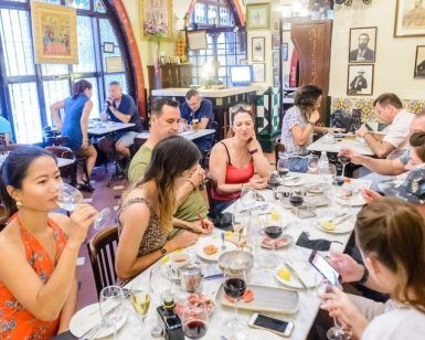 Gente disfrutando de un "Tour de tapas por Barcelona" en un animado restaurante, cenando en una mesa larga con bebidas, rodeados de una decoración colorida que recuerda a una vibrante noche de Barcelona. - ForTwo - Planes para parejas