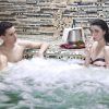 Dos personas se relajan en el jacuzzi de su patio trasero; una disfruta de una copa con una botella de vino en una nevera cercana, evocando el encanto de Sevilla. Es como vivir su propia experiencia del Circuito de Spa en las Termas de Hispalis en casa. - ForTwo - Planes para parejas