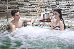 Dos personas se relajan en el jacuzzi de su patio trasero; una disfruta de una copa con una botella de vino en una nevera cercana, evocando el encanto de Sevilla. Es como vivir su propia experiencia del Circuito de Spa en las Termas de Hispalis en casa. - ForTwo - Planes para parejas